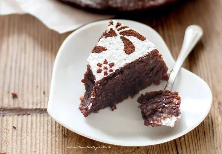 Una fetta di torta caprese ricetta originale napoletana, cosparsa di zucchero a velo e cacao, su un piatto bianco a forma di cuore.