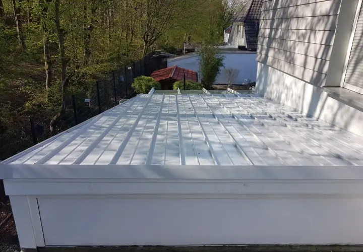 Neues, weißes Metalldach mit sichtbarem Ortgang. Im Hintergrund ein kleiner Garten mit Bäumen und einem roten Gartenhaus.