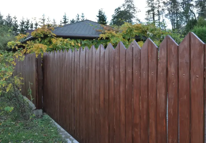 Brązowe, drewniane sztachety z ostrymi zakończeniami tworzą płot. Widać, ile sztachet na metr jest potrzebne do zbudowania takiego ogrodzenia.