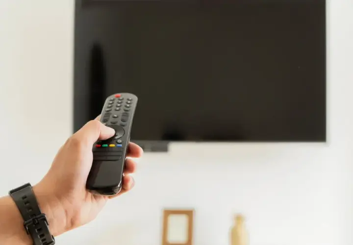Dłoń trzymająca pilot do telewizora na tle wyłączonego ekranu.