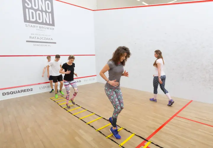Grupa osób wykonuje dynamiczny stretching na drabince gimnastycznej w sali do squasha.