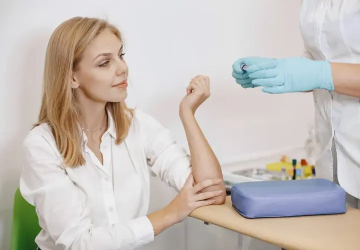 Młoda kobieta z blond włosami czeka na pobranie krwi. Pielęgniarka w niebieskich rękawiczkach trzyma probówkę. Badanie na czczo.