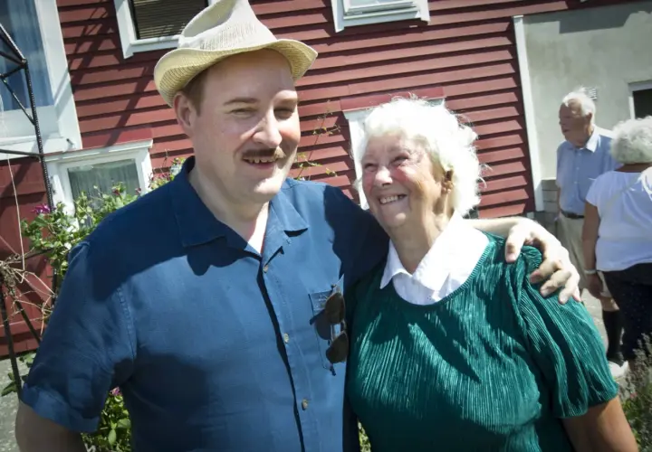 Sigge Dorsin ler mot kameran med en äldre kvinna. De står framför ett rött hus.