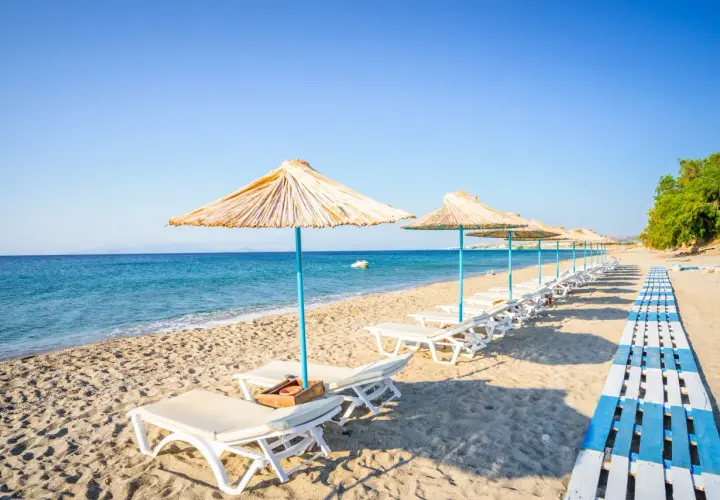 Słoneczna plaża na wyspie Kos, rzędy leżaków pod słomianymi parasolami, turkusowe morze i błękitne niebo. Idealne miejsce na relaks.