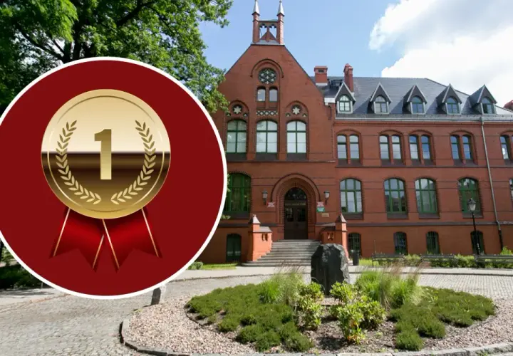 Złoty medal z cyfrą "1" na tle ceglanego budynku i zieleni.