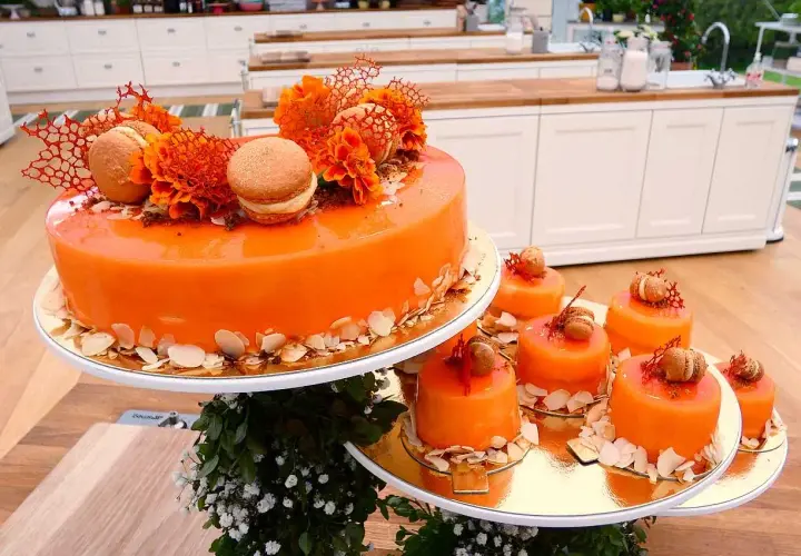 En stor och flera små orangea tårtor med spegelblank glasyr, dekorerade med makroner och blommor.
