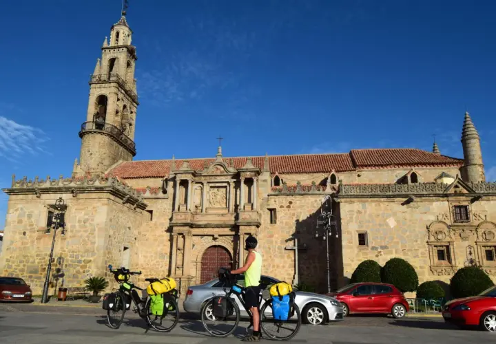 Ciclistas preparan sus bicicletas para continuar las etapas de Transandalus frente a una iglesia histórica bajo un cielo azul.