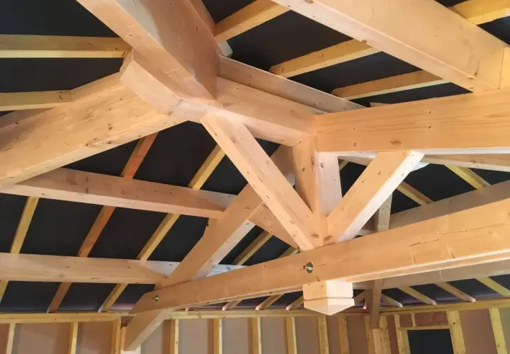 Vue rapprochée d'une charpente traditionnelle 2 pans en bois clair, avec des poutres massives et des chevrons assemblés.