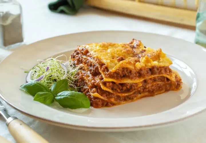 Porción de lasaña de carne casera, con capas de pasta, salsa boloñesa y queso gratinado, servida con brotes y albahaca.