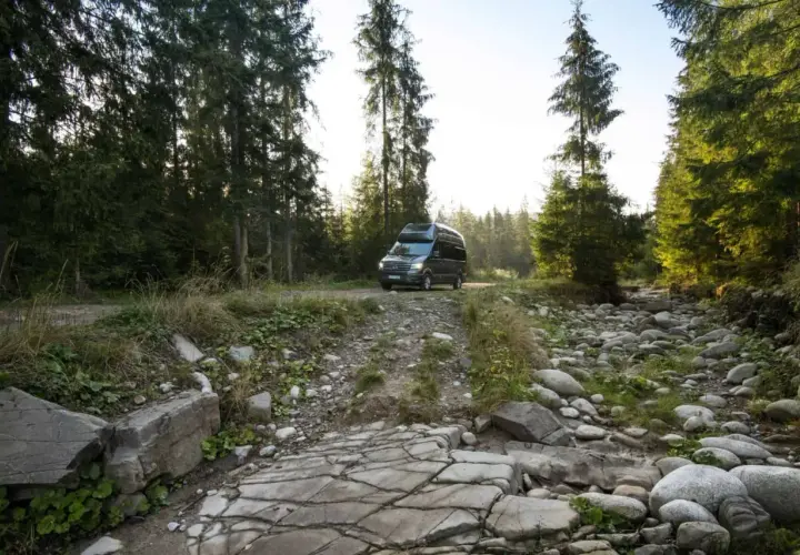 Ciemny kamper na leśnej drodze, otoczony przez wysokie drzewa i kamieniste koryto strumienia. Idealne miejsce na **split camping** i ucieczkę od cywilizacji.