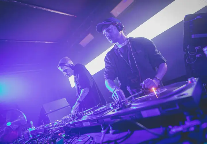 Dwóch DJ-ów w słuchawkach tworzy muzykę techno. Jeden z nich ma na szyi identyfikator "ARTIST".
