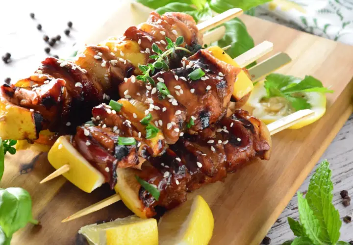 Aromatyczne yakitori z kurczaka w sosie teriyaki, posypane sezamem i szczypiorkiem, podane z cytryną na drewnianej desce.