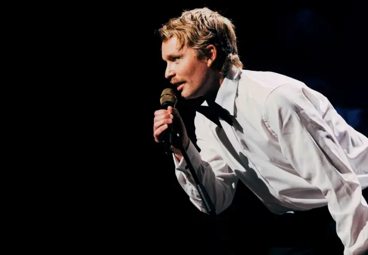 En man med mustasch, klädd i vit skjorta, sjunger i mikrofonen under nalles show.
