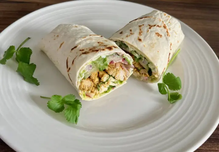 Kebab w tortilli przekrojony na pół, z kurczakiem, warzywami i ziołami, na białym talerzu.