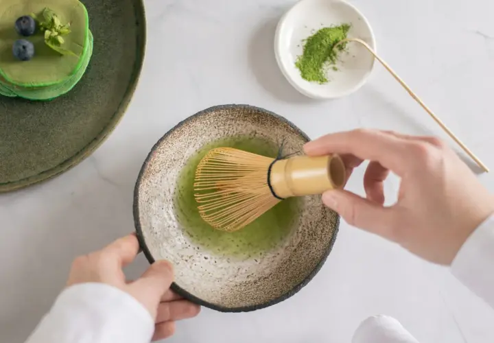 Hände bereiten Matcha zu, ein gesunder Genuss für die matcha schwangerschaft. Daneben grüne Pancakes mit Blaubeeren.