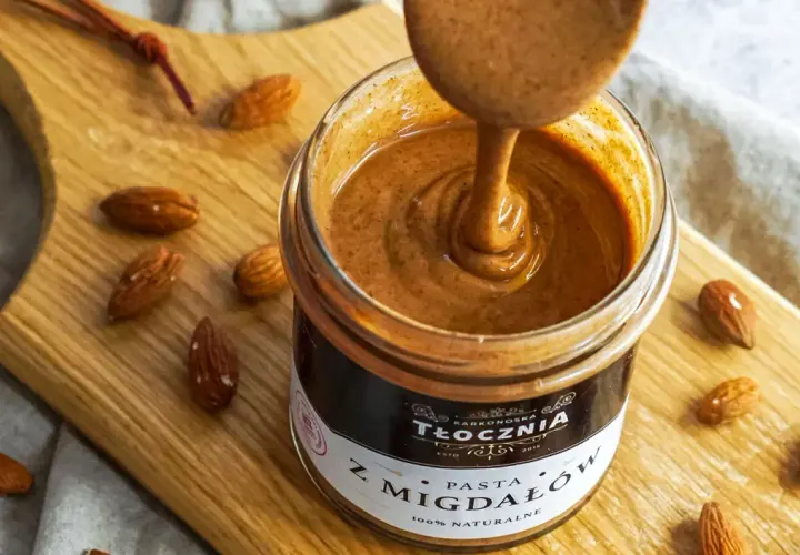 Gęsta, kremowa pasta z migdałów, czyli polskie "noisette", w słoiku, otoczona migdałami na drewnianej desce.
