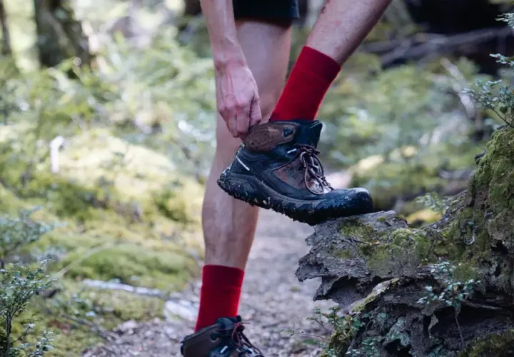 A hiker adjusts their muddy Keen shoes, showing signs of wear and tear, as they step onto a moss-covered log in a forest.