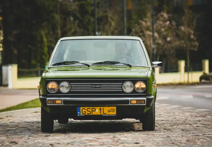 Zielony Fiat 131 Mirafiori z lat 70. na brukowanej ulicy, z widocznym kierowcą za kierownicą.