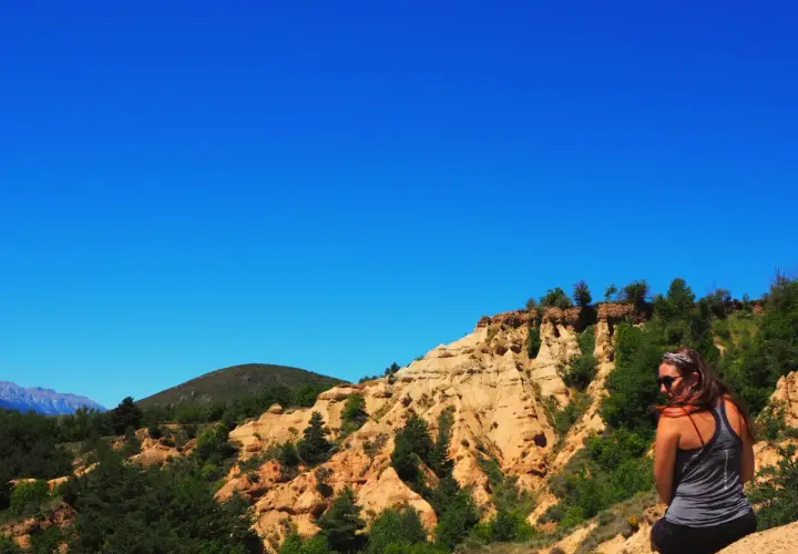 Una mujer contempla el paisaje de la Cerdanya, con sus formaciones rocosas ocres y vegetación verde bajo un cielo azul intenso.