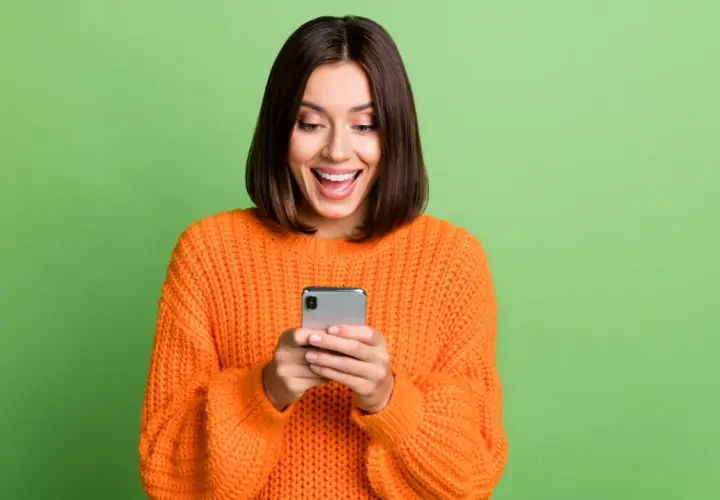 Uśmiechnięta kobieta w pomarańczowym swetrze trzyma smartfon. Może właśnie dostała informację o wyłączeniu 3G Orange i cieszy się z nowej oferty.