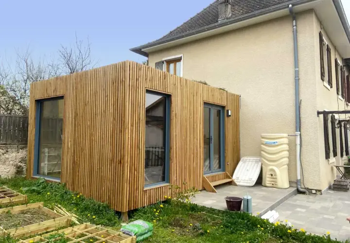 Une extension en bois moderne s'intègre à une maison existante, avec de grandes fenêtres et une finition en lattes de bois.