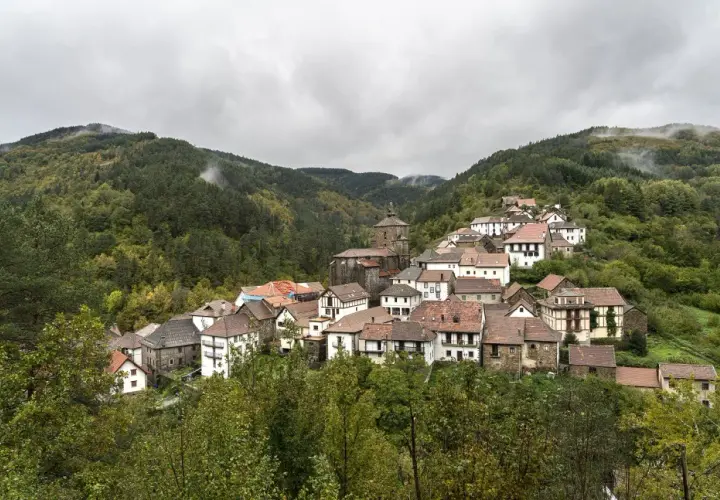 Pueblo pintoresco en el Pirineo Navarro, con casas de piedra y tejados rojos, rodeado de bosques frondosos y montañas nubladas.