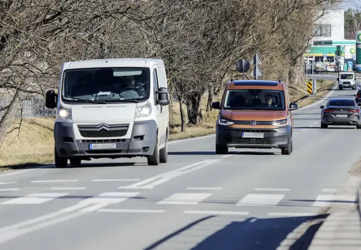 Biała furgonetka wyprzedza na przejściu dla pieszych pomarańczowego vana. W tle stacja benzynowa.