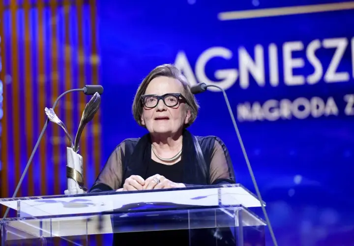 Agnieszka Holland w okularach za mównicą z mikrofonem i statuetką.