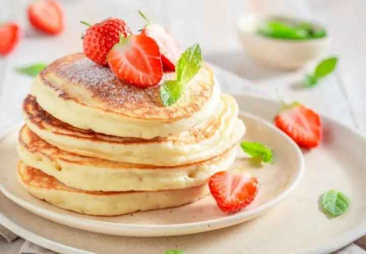 Puszyste ciasto na pancake, ozdobione świeżymi truskawkami i listkami mięty, wygląda apetycznie.