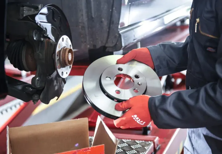 Mechanik w czerwonych rękawiczkach trzyma nową tarczę hamulcową obok zamontowanej tarczy.