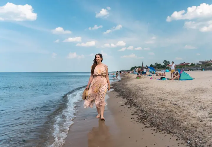 Kobieta w kwiecistej sukience spaceruje brzegiem morza. W tle plaża pełna ludzi, parawanów i domków – typowe sarbinowo atrakcje.