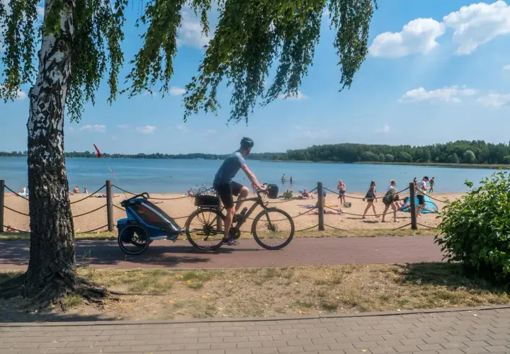 Rowerzysta z przyczepką jedzie ścieżką przy plaży nad Pogorią. Ludzie odpoczywają na piasku.