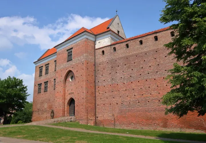 Zamek w Łęczycy, ceglana twierdza z czerwonej cegły, z charakterystycznym łukowym wejściem i oknami, otoczona zielenią.