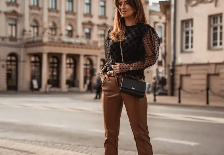 Elegancka kobieta w brązowych spodniach i czarnej bluzce w kropki, z czarną torebką na łańcuszku, pozuje na tle miejskiej kamienicy.