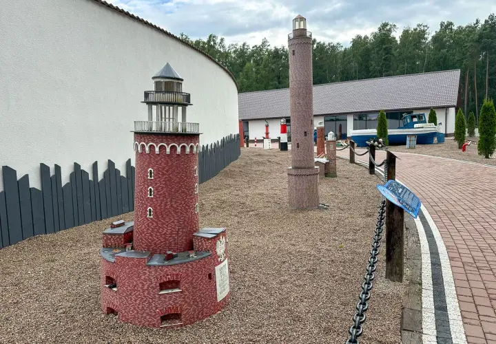 Miniaturowa latarnia morska z czerwonej cegły, obok wyższa, kamienna. W tle budynek i łódź.