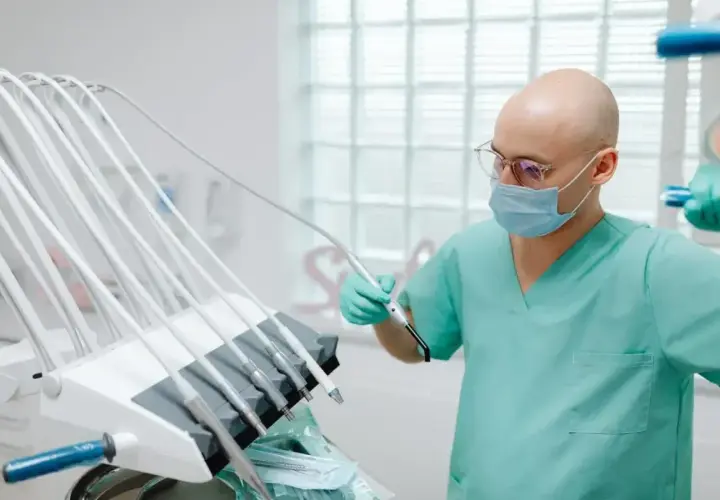 Dentysta w masce i okularach trzymający narzędzie przy unitu stomatologicznego.