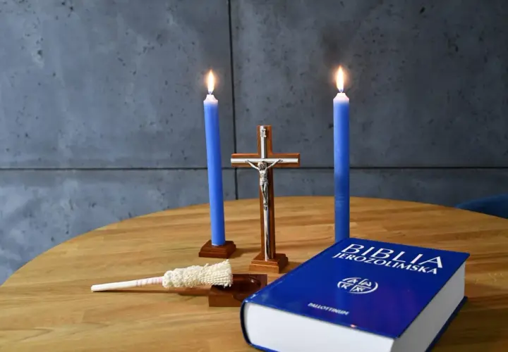 Dwie niebieskie świece, krzyż, kropidło i Biblia Jerozolimska na drewnianym stole. Symbolika religijna, jakby przygotowanie do kolędy.