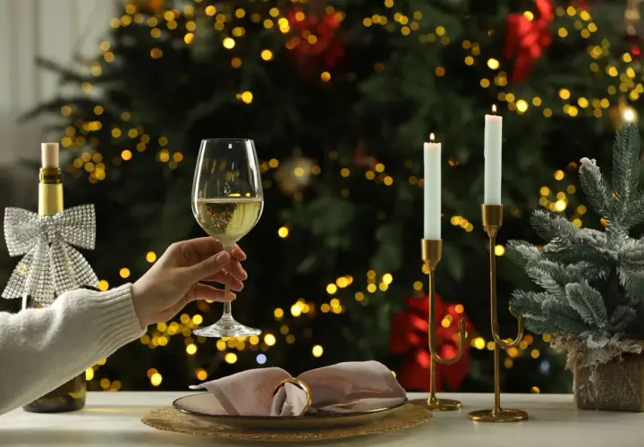 Une main tient un verre de vin blanc pétillant, parfait pour un buffet froid. Une bouteille décorée et des bougies illuminent la scène devant un sapin de Noël scintillant.