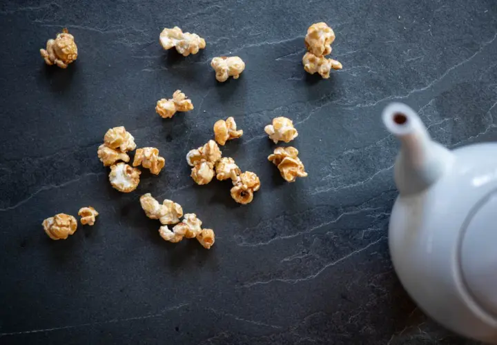 Pop corn caramel éparpillé sur une surface sombre, avec une théière blanche en arrière-plan.