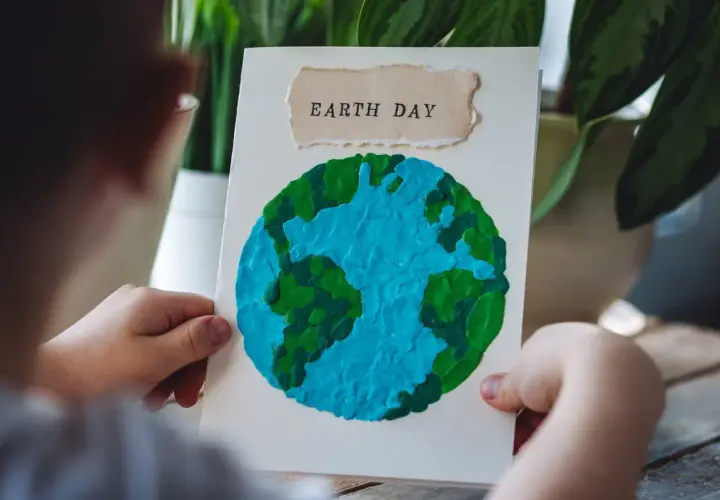Dziecko trzyma kartkę z pracą plastyczną przedstawiającą planetę Ziemię z plasteliny. Napis "EARTH DAY" na górze.