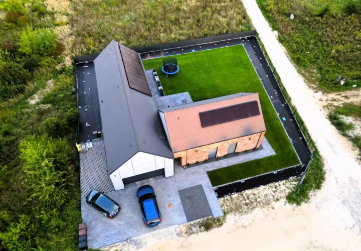 Widok z lotu ptaka na nowoczesny dom z panelami słonecznymi, ogrodem i samochodami.