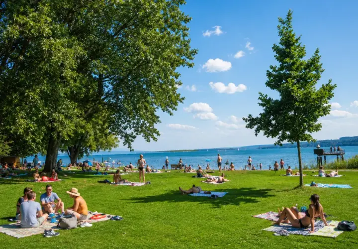 Sonniger Tag im Strandbad Bodensee. Menschen entspannen auf Decken im Gras, baden im See und genießen die Aussicht.