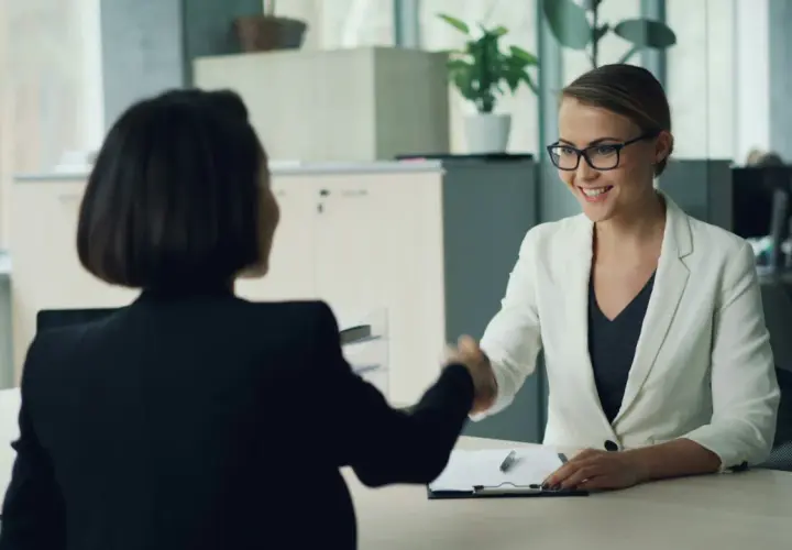 Zwei Frauen schütteln sich die Hände. Eine Frau im weißen Blazer lächelt, während sie eine Bewerbung für personalentwicklung jobs prüft.