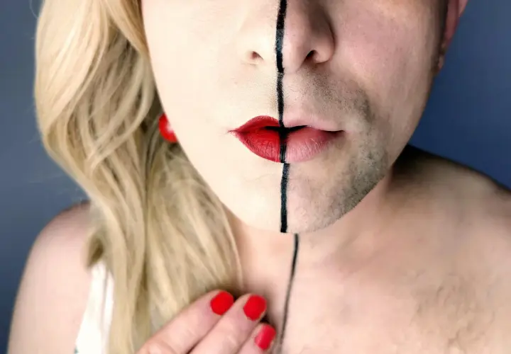 Un volto diviso a metà, con capelli biondi e barba, labbra rosse e una linea nera. Un'espressione di una persona non binaria che abbraccia la propria identità.