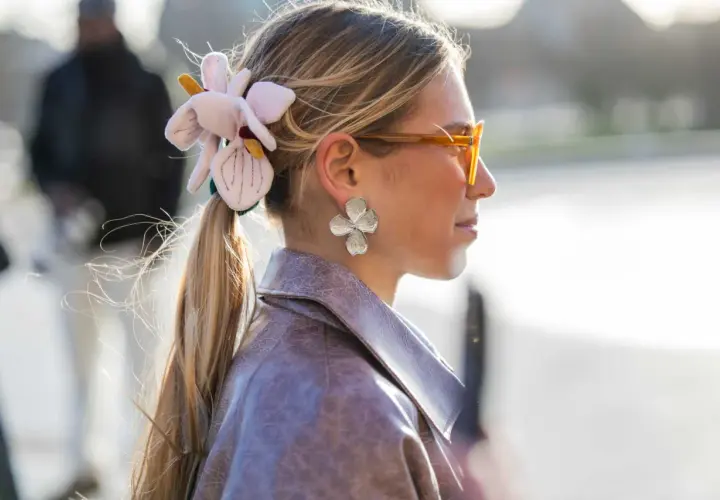 Mujer con coleta adornada con una flor de tela y pendientes de pétalos, luciendo gafas naranjas y una chaqueta de cuero.