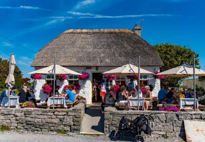 Café pittoresque avec toit de chaume, parasols et fleurs. Des gens dégustent un repas en plein air, peut-être un délicieux aran poisson.