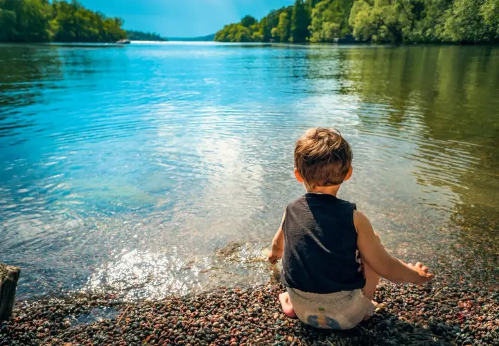 Un bambino seduto sulla riva di un lago, godendosi un tranquillo week end con bambini. L'acqua limpida riflette il cielo azzurro e gli alberi verdi.