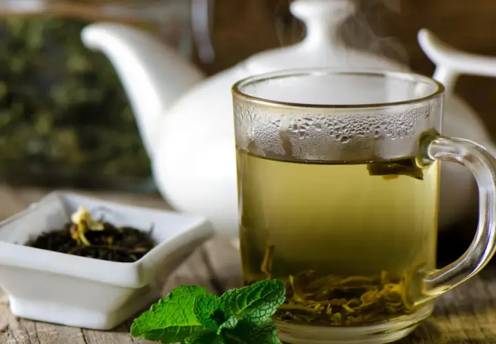 Dampfender grüner Tee in einer Tasse, daneben eine Schale mit Teeblättern und Minze. Welcher Tee ist antibakteriell? Grüner Tee ist bekannt für seine gesundheitlichen Vorteile.
