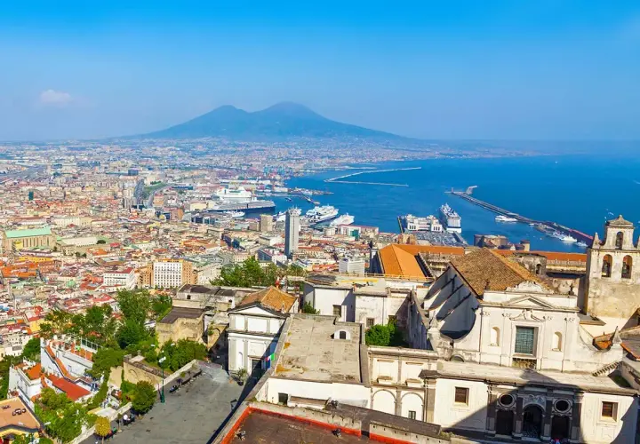 One day in Naples, a panoramic view reveals the city's sprawl, the bustling port with cruise ships, and the majestic Mount Vesuvius in the distance under a clear blue sky.