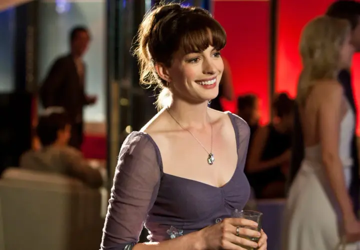 Uśmiechnięta Anne Hathaway w fioletowej sukience, trzymająca drinka. Scena z jednego z jej filmów.